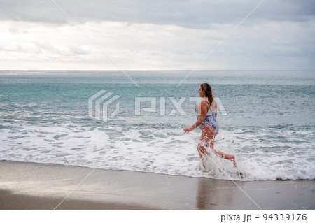 A plump woman in a bathing suit enters the water during the surf. Alone on the beach, Gray sky in the clouds, swimming in winter. 94339176