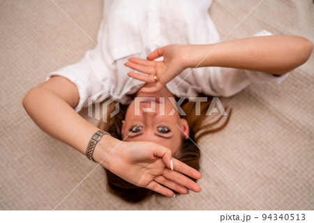 The woman lies on the bed on her back, top view. She looks straight ahead, wearing a white shirt 94340513