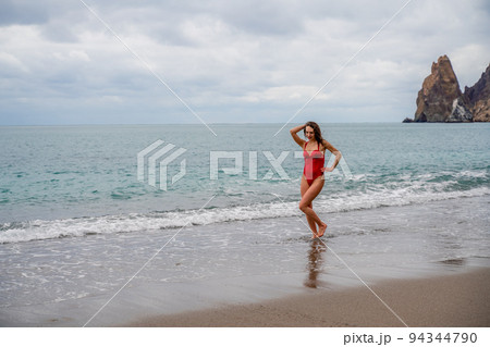 A beautiful and sexy brunette in a red swimsuit on a pebble beach, Running along the shore in the foam of the waves 94344790