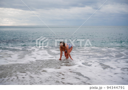 A plump woman in a bathing suit enters the water during the surf. Alone on the beach, Gray sky in the clouds, swimming in winter. 94344791