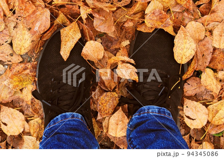 Women's legs in blue jeans and black sneakers against the background of autumn yellow-orange leaves. View from above. 94345086