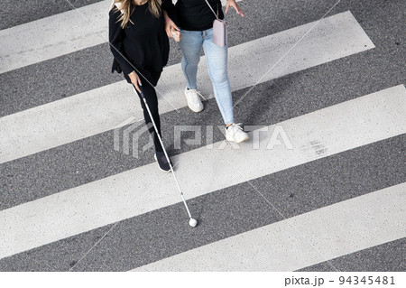 Scene of a Blind woman walking on zebra crossing helped by another person using her white cane. Help in the early stages of blindness Scene of a Blind woman walking on zebra crossing helped by another person using her white cane. Help in the early stages of blindness 94345481