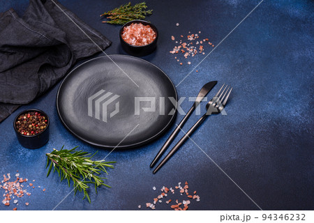 An empty black plate on a dark concrete background with spices and herbs 94346232