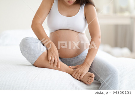 Pregnant woman massaging her swollen foot, sitting on bed 94350155