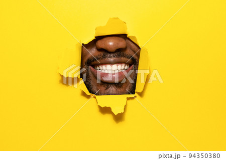 Perfect smile. African american man smiling and showing teeth through torn bright yellow color paper background 94350380