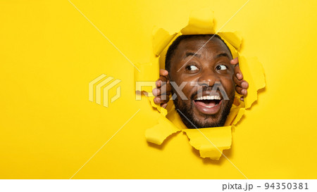 Excited african american man smiling and looking away while tearing hole in yellow paper background, copy space Excited african american man smiling and looking away while tearing hole in yellow paper background, copy space 94350381