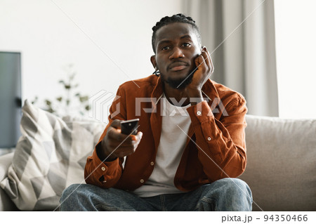 Boring television program. Bored upset black man watching TV and switching channels with remote control, sitting on sofa Boring television program. Bored upset black man watching TV and switching channels with remote control, sitting on sofa 94350466