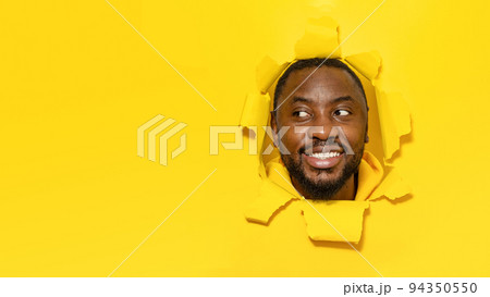 Happy black man looking aside at free space and smiling, noticing something interesting, yellow paper hole 94350550