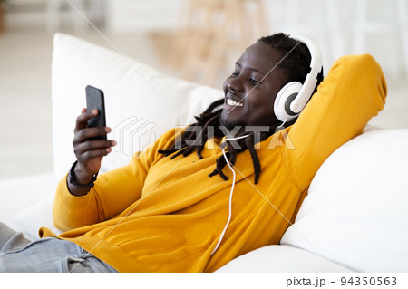 Portrait Of Smiling Black Man Wearing Headphones Relaxing With Smartphone At Home Portrait Of Smiling Black Man Wearing Headphones Relaxing With Smartphone At Home 94350563