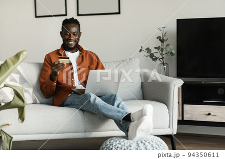 Excited african american man sitting on couch at home using laptop choosing what to buy and holding credit card 94350611