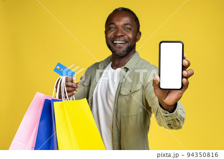 Smiling mature african american guy in casual show smartphone with empty screen and many shopping bags Smiling mature african american guy in casual show smartphone with empty screen and many shopping bags 94350816