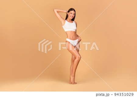 Fit young lady with smooth skin and perfect body shape posing in white lingerie over beige background, full length Fit young lady with smooth skin and perfect body shape posing in white lingerie over beige background, full length 94351029