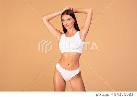 Portrait of fit woman in white lingerie posing on beige studio background, raising arms up, showing smooth armpits Portrait of fit woman in white lingerie posing on beige studio background, raising arms up, showing smooth armpits 94351032