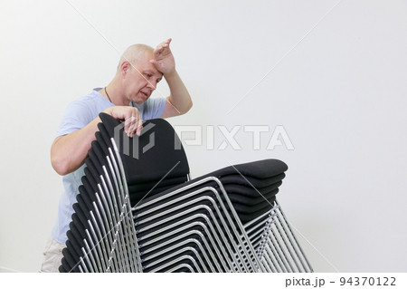 tired gray-haired man wipes sweat from his forehead.he pushes a stack of chairs  94370122