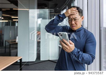 Asian businessman inside office building upset and sad reading bad news on mobile phone, business owner investor using smartphone standing near window 94373587