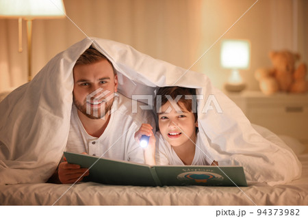 Father and son with flashlight reading book under blanket at home Father and son with flashlight reading book under blanket at home 94373982