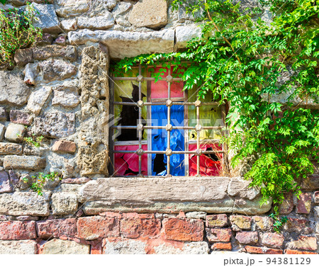 Weathered old stone bricks wall with corroded antique grunge wrought iron window with multi vibrant colored squares Weathered old stone bricks wall with corroded antique grunge wrought iron window with multi vibrant colored squares 94381129