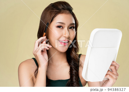 Portrait of ardent female model applying her eyeshadow and holding a mirror. Portrait of ardent female model applying her eyeshadow and holding a mirror. 94386327