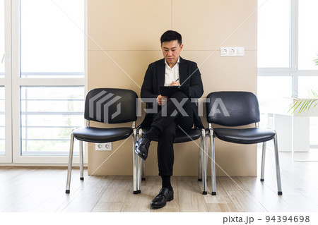 Young successful Asian man, waiting for a job interview, in the waiting room of the office center, sitting on a chair near the reception of the office center Young successful Asian man, waiting for a job interview, in the waiting room of the office center, sitting on a chair near the reception of the office center 94394698