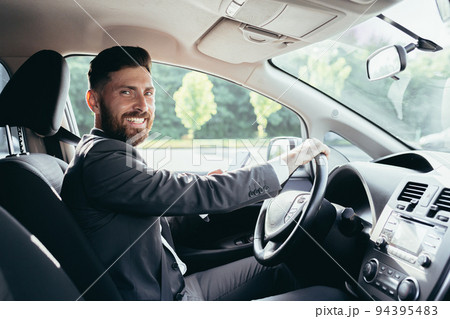 Young man with a beard businessman driving a car in the parking lot, looking at the camera and smiling 94395483