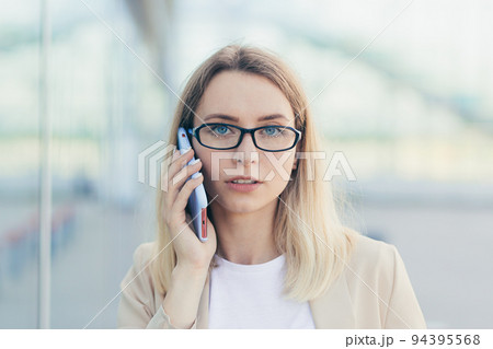 portrait of a serious woman wearing glasses blonde holding a mobile phone talking on a smartphone near the office 94395568