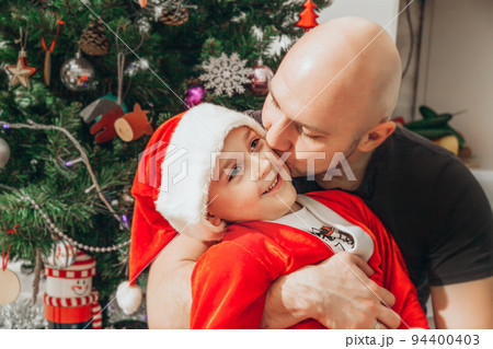 Dad kisses his son, dressed in a Santa costume, near the Christmas tree 94400403