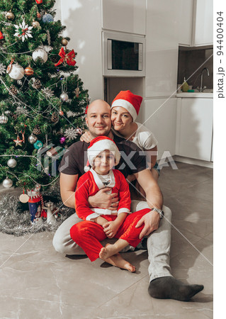 Family - mom, dad and son in Santa costume sitting under the Christmas tree on the floor in their new apartment, looking into the camera Family - mom, dad and son in Santa costume sitting under the Christmas tree on the floor in their new apartment, looking into the camera 94400404