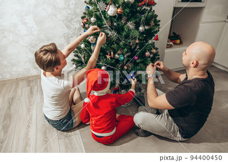 Family - mom, dad and son in a Santa costume are sitting under the Christmas tree on the floor in their apartment and decorating the Christmas tree Family - mom, dad and son in a Santa costume are sitting under the Christmas tree on the floor in their apartment and decorating the Christmas tree 94400450