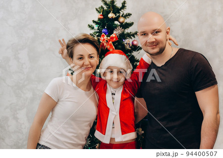 Family mom, dad and son in Santa costume are standing near the Christmas tree in their apartment and looking into the camera 94400507