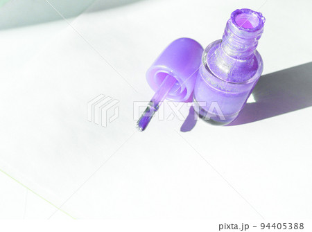 An open bottle with lilac nail polish and a brush next to it on a white surface with a shadow, the concept of fashionable beauty 94405388