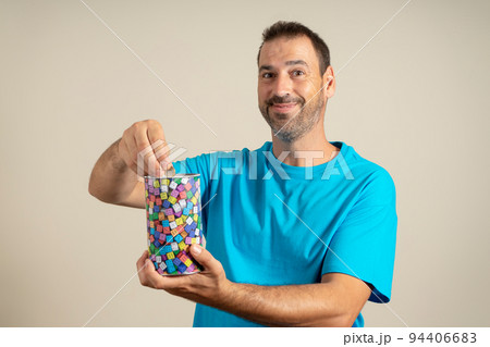 Smiling latin man with a beard dressed in a blue t-shirt putting a euro coin in a metal piggy bank, trying to save so he can travel on vacation. Isolated on beige studio background 94406683