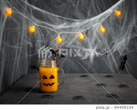 Halloween pumpkin juice in a glass jar with a scary face, spiders and cobwebs on a dark gray background. Funny decorations for a children's party. Copy space. Halloween pumpkin juice in a glass jar with a scary face, spiders and cobwebs on a dark gray background. Funny decorations for a children's party. Copy space. 94409104