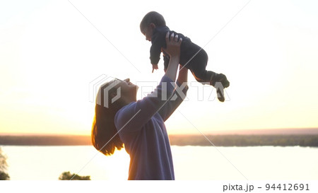 A young mother holds her baby son in her arms at sunset. A woman with glasses on a hill raises a child up against the background of the river. Close-up. A young mother holds her baby son in her arms at sunset. A woman with glasses on a hill raises a child up against the background of the river. Close-up. 94412691