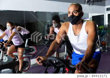 Men and women wearing protective masks ride stationary bike in fitness club 94413171