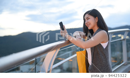 Smiling young woman typing text message on mobile phone while standing on roof terrace outdoors with cityscape on background Smiling young woman typing text message on mobile phone while standing on roof terrace outdoors with cityscape on background 94425322
