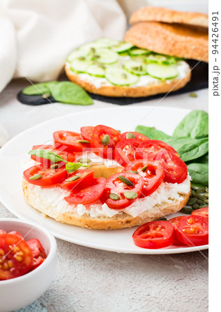 Delicious bagel stuffed with feta, tomatoes and pumpkin seeds and spinach leaves on a plate. Light Healthy Vitamin Snack. Vertical view. Close-up Delicious bagel stuffed with feta, tomatoes and pumpkin seeds and spinach leaves on a plate. Light Healthy Vitamin Snack. Vertical view. Close-up 94426491