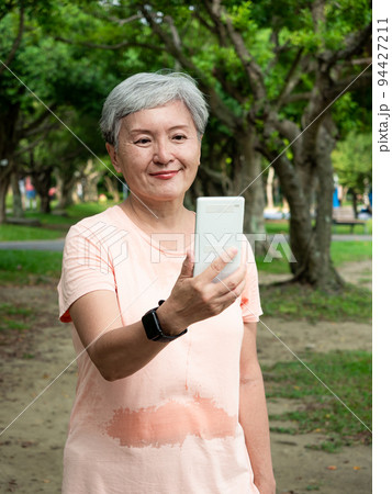 Portrait of senior adult elderly asian woman 60s smiling standing and holding smartphone in the park. 94427211