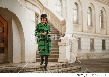 Outdoor fashion portrait of an elegant fashionable brunette woman, model in a stylish cap, green dress, posing at sunset in a European city in autumn. 94428204