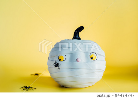 Funny and cute pumpkin mummy with spider on the yellow background. White Pumpkin with scaried and worried look. Fear of insects. Halloween decor and background with copy space. Selective focus 94434747