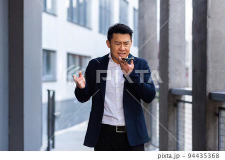 angry and serious Successful Asian businessman explains information to employees using phone, speaks near office outside angry and serious Successful Asian businessman explains information to employees using phone, speaks near office outside 94435368