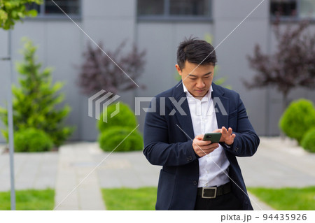 Asian businessman boss looks at mobile phone reads news from phone and writes in messenger outside office Asian businessman boss looks at mobile phone reads news from phone and writes in messenger outside office 94435926