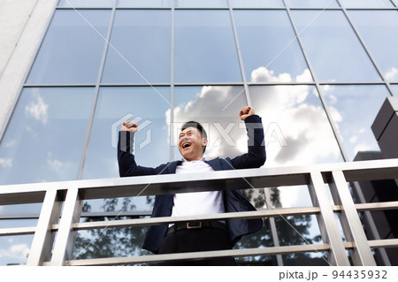 Asian businessman boss stands near his office center on the balcony in a dark color business suit holds his hands up celebrates victory Asian businessman boss stands near his office center on the balcony in a dark color business suit holds his hands up celebrates victory 94435932