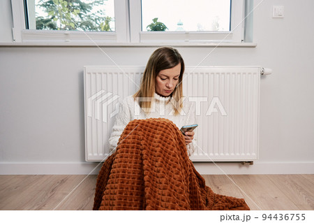 Worried sad woman sits under blanket near heating radiator and use smartphone, Rising costs in private households for gas bill due to inflation and war, Energy crisis 94436755