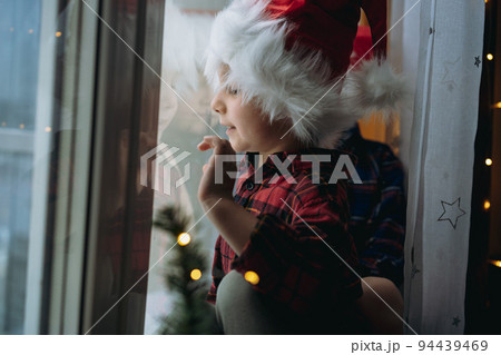cute caucasian child wearing santa hat and plaid shirt sitting hugging by the window waiting for christmas 94439469