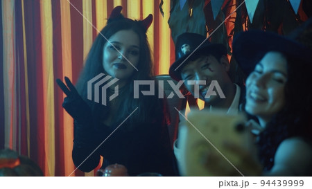 A group of friends are sitting at a table in creepy costumes, taking pictures with a camera and laughing on Halloween 94439999