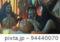 Two girls and a guy in creepy Halloween costumes are sitting at a table chatting and carving faces on pumpkins 94440070