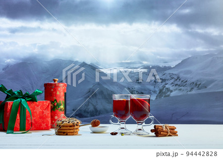 Hot mulled wine in glasses with orange slices, cinnamon sticks and spices with Christmas decorations on table over background of landscape of winter snow-capped mountains Hot mulled wine in glasses with orange slices, cinnamon sticks and spices with Christmas decorations on table over background of landscape of winter snow-capped mountains 94442828