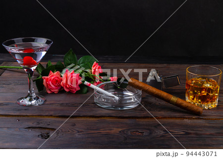 Alcoholic cocktails for man and woman on wooden desk. Glass of whiskey with a cigar on wooden table. Bouquet of flowers and cigarette in ashtray. 94443071