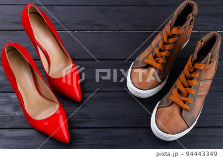 Red female high heel shoes and brown male sneakers. Top view mens and womens shoes on dark wooden background. Red female high heel shoes and brown male sneakers. Top view mens and womens shoes on dark wooden background. 94443349