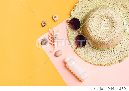 Beach vacation. A straw hat, sunglasses, a bottle of sunscreen and shells on a yellow-pink cardboard. Items for sunbathing and tanning. Top view Beach vacation. A straw hat, sunglasses, a bottle of sunscreen and shells on a yellow-pink cardboard. Items for sunbathing and tanning. Top view 94444870